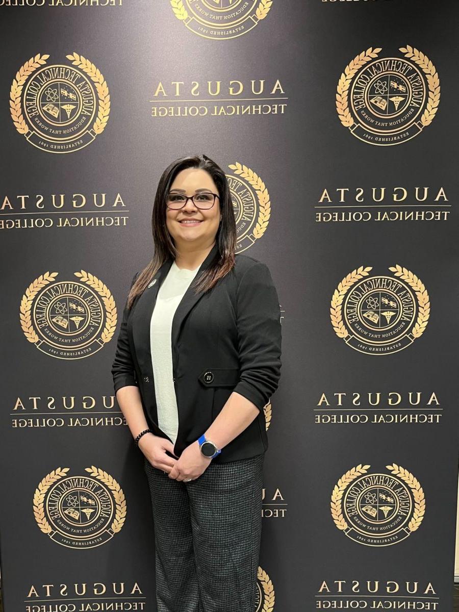 Devlynn Taylor, a caucasian female, stands in front of a backdrop with the Augusta Technical College seal and name in black and gold. She is wearing a black blazer with a white crew neck dress shirt and dark grey dress pants.